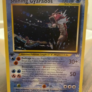 Shining Gyarados Pokémonkort - Säljer ett Shining Gyarados Pokémonkort med blå ram och holografisk bild. Kortet har 100 HP och visar Gyarados i en dramatisk scen med båt och vatten. Text och attacker på engelska. Perfekt för samlare av sällsynta Pokémonkort.