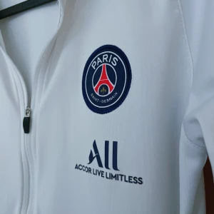 PSG Fotbollströja Nike - Säljer en vit träningsjacka från Nike med PSG-logga på bröstet och röd Nike-swoosh på ärmarna. Tröjan har dragkedja framtill och är långärmad. Perfekt för fotbollsträning eller som en sportig vardagsjacka.