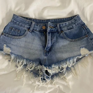Blå jeansshorts med fransar - Säljer ett par blå jeansshorts med slitna detaljer och fransig kant. Shortsen har klassisk femficksdesign, låg midja och råa kanter för en trendig look. Perfekta för sommaren och festivaler.🌸