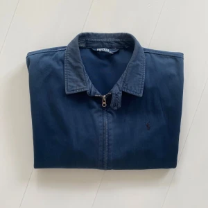 Blå jacka från Polo Ralph Lauren - Säljer en klassisk blå jacka från Polo Ralph Lauren med dragkedja framtill och diskret broderad logga på bröstet. Jackan har krage, två snedställda fickor och långa ärmar med knapp vid ärmslut. Perfekt för vår, sommar och höst. Boxyfit, size XL men sitter mer som L. Den eftertraktade modellen utan foder i 100% bomull.