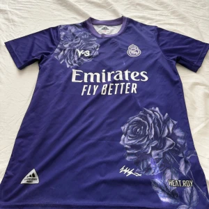 Real Madrid x Adidas Y-3 lila fotbollströja - Säljer en lila Real Madrid fotbollströja från Adidas Y-3-kollektionen med stora blommönster, klubbmärke och sponsortryck på bröstet. VINI JR och nummer 7 på ryggen. Kortärmad modell i lätt funktionsmaterial.