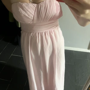 Ljusrosa balklänning  - Säljer ljusrosa långklänning med axelbandslös design och vackert draperad överdel. Klänningen har markerad midja och faller mjukt ner mot golvet. Perfekt för bal eller festliga tillfällen.