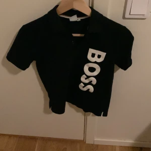 Svart piké från BOSS med tryck - Säljer en piké från BOSS med stort vitt tryck på framsidan. Piken har krage och knappar upptill för en stilren look. Perfekt för dig som gillar klassiska märken och enkel design. Du ansvarar för frakt.