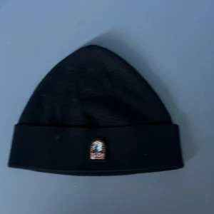 Svart mössa från Parajumpers - Svart ribbstickad mössa från Parajumpers med uppvikt kant och liten broderad logotyp framtill. Klassisk och stilren modell som passar perfekt till kyliga dagar. Storlek L-Xl knappast använd. 