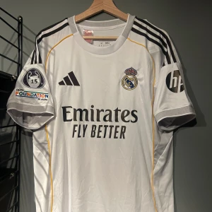 Real Madrid fotbollströja Bellingham 5 - Säljer en vit Real Madrid matchtröja med Bellingham och nummer 5 på ryggen. Tröjan har svarta detaljer, klubbmärke på bröstet, Champions League-märke på ärmen och sponsorloggor. Klassisk design med korta ärmar och gula inslag längs sidorna. Helt ny en replika med tagg