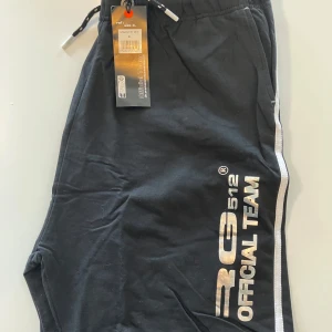 Svarta shorts med vit rand och tryck från RG512 - Svarta shorts från RG512 med vit rand längs sidorna och stort silvrigt tryck 'RG512 OFFICIAL TEAM' på ena benet. Resår i midjan med snörning och två sidofickor. Perfekta för en sportig och avslappnad stil.
