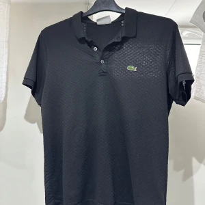 Svart pikétröja från Lacoste - Klassisk svart pikétröja från Lacoste med korta ärmar och krage. Tröjan har två knappar framtill och den ikoniska gröna krokodilloggan på bröstet. Perfekt för en stilren och sportig look.