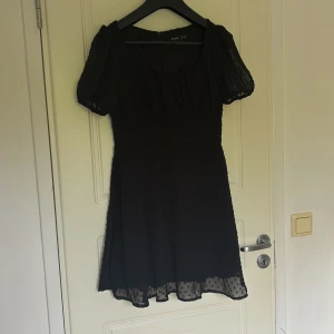 Svart kortklänning med puffärm från SHEIN - Svart kortklänning från SHEIN med puffärmar och diskret prickigt mönster i tyget. Klänningen har rundad halsringning och lätt utställd kjol. Perfekt för dig som gillar en feminin och trendig stil.