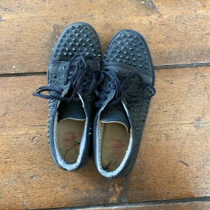Svarta sneakers med nitar från Christian Louboutin - Säljer ett par svarta sneakers från Christian Louboutin med svarta nitar över hela skon och klassisk röd sula. Skorna har snörning. Perfekta för dig som vill sticka ut med en edgy stil. En nit saknas dock på sidan av skon kan gå ner i pris vid snabb affär