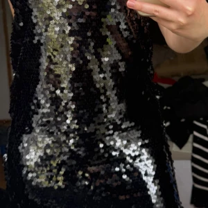 Svart paljettklänning - Säljer en svart axelbandslös klänning täckt av glittrande paljetter. Klänningen har en lösare passform och är perfekt för dig som vill sticka ut. Supersnygg till fest och nyår! Lappen sitter kvar på klänningen och har aldrig använts.