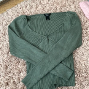 Grön ribbad långärmad topp från LinDEX - Snygg grön långärmad topp från LinDEX i ribbat material. Toppen har en v-ringad hals och en liten knytdetalj framtill. Perfekt att styla med jeans eller kjol för en trendig look. Framtill är den fäst med en säkerhetsnål väldigt öppen 
