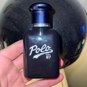 Polo Ralph Lauren parfymflaska - Mörkblå parfymflaska från Polo Ralph Lauren med vit logotyp och text 'EST. 67' på framsidan. Flaskan har rundad form och metallring vid halsen med ingraverad text. Perfekt för dig som gillar manliga och fräscha dofter. Perfekt till alla årstider! Ungefär 25ml kvar 