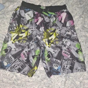 Färgglada badshorts från Kappa - Säljer ett par badshorts från Kappa med coolt collage-mönster i svart, grått, gult, grönt, blått och rosa. Shortsen har elastisk midja med grön snörning och innerbyxa i mesh. Perfekta för stranden eller poolen!