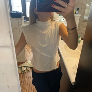 Vit ärmlös topp med knyt - Säljer en vit ärmlös topp med hög hals och knytdetalj i midjan. Toppen har en stilren och modern design som passar perfekt till många olika outfits.