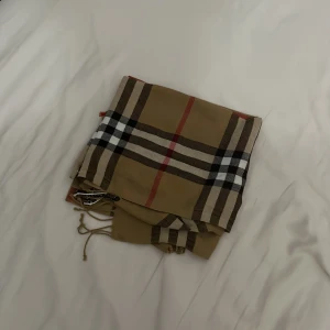 Beige rutig halsduk från Burberry - Säljer en klassisk beige halsduk från Burberry med det ikoniska rutiga mönstret i svart, vitt och rött. Halsduken har broderad Burberry-logga och fransar i kanterna. Perfekt accessoar för att lyfta din outfit.