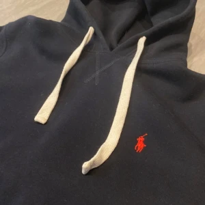 Mörkblå hoodie från Polo Ralph Lauren - Säljer en mörkblå hoodie från Polo Ralph Lauren med vit dragsko och röd broderad logga på bröstet. Klassisk känguruficka framtill och mjukt material som känns skönt mot huden.