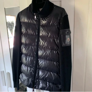 Svart dunväst från Moncler med stickade ärmar - Säljer en svart dunväst från Moncler med glansig yta och stickade svarta ärmar. Västen/Jackan har dragkedja framtill och en ficka med Moncler-logga på ena ärmen. Perfekt för lager-på-lager-stil. Kvitto finns! Köpt på Monclers butik i Stockholm.