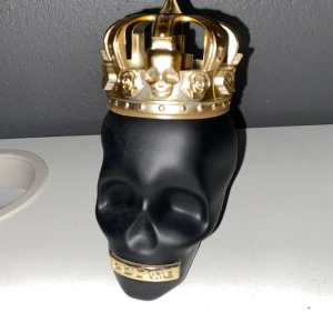 Black Skull parfymflaska med guldkrona - Unik parfymflaska från Police formad som ett svart dödskallehuvud med en detaljerad guldfärgad krona på toppen. Flaskan har även en guldfärgad detalj vid munnen med text. Perfekt för dig som gillar cool och annorlunda design. Flaskan är 125 ml så uppskattar att det är iaf mer än halva kvar, skulle tippa 90-100 ml!