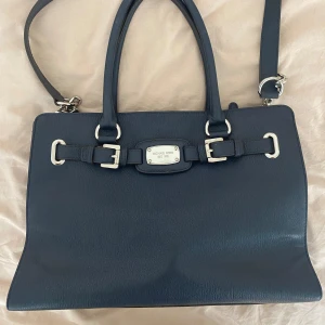 Mörkblå handväska från Michael Kors - Snygg mörkblå handväska från Michael Kors med silverfärgade metalldetaljer och dekorativa spännen framtill. Väskan har både handtag och axelrem samt flera fack inuti. Använd ett fåtal gånger.