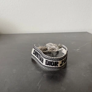 Vit armband från Dior - Säljer ett snyggt vävt armband från Dior i beige och svart med texten 'Christian Dior J'Adior'. Armbandet har justerbar knytning med tofsar i ändarna. Perfekt accessoar för att lyfta din outfit.