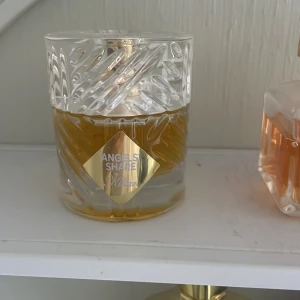 Angels' Share Eau de Parfum från Kilian - Exklusiv parfym från Kilian, Angels' Share, i en lyxig glasflaska med snidad design och guldfärgad etikett. Doften är en Eau de Parfum och flaskan har en elegant, rund form med genomskinligt glas och gyllene vätska.