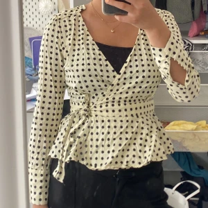 Prickig omlottblus med knyt och volang - Snygg vit omlottblus med svarta prickar och knyt i midjan. Blusen har långa ärmar med knappar vid slutet av ärmen💕💕