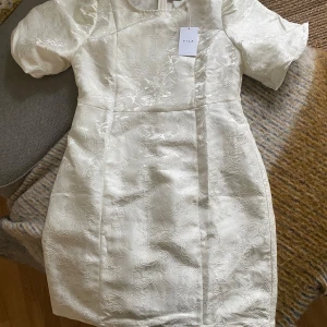 Vit kortklänning med blommigt mönster från Vila - Säljer en elegant vit kortklänning från Vila med subtilt blommigt mönster. Klänningen har korta puffärmar och en figurnära passform. Perfekt för sommarens tillfällen och studentfirande. Materialet har en lätt glans och struktur. Helt oanvänd med prislapp kvar.