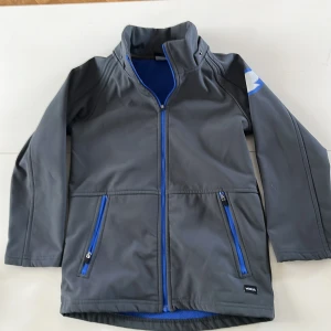 Grå softshell regnjacka från MORXS med blå detaljer - Grå softshelljacka från MORXS med blå dragkedjor och blått fleecefoder. Jackan har hög krage, två fickor med dragkedja och lång ärm. Perfekt för kyliga dagar och har en snygg detalj på ärmen. Tillhörande luva finns inte kvar.  Sparsamt använd. 