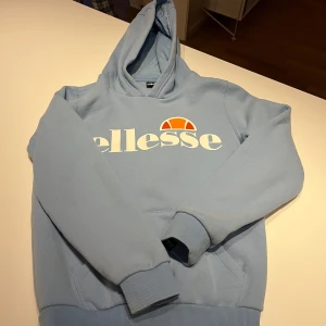 Ljusblå hoodie från Ellesse 12/13år  - Säljer en ljusblå hoodie från Ellesse med stor vit logga och orange detalj på bröstet. Tröjan har huva och magficka, samt ribbade muddar vid ärmslut och nederkant. 