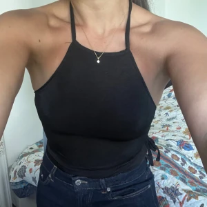 Svart croppad halterneck-linnetopp med knytband - Säljer en svart croppad halterneck-linnetopp med smala axelband och knytning i sidorna. Toppen är ärmlös och har öppen rygg, perfekt för varma dagar eller en snygg layering-look.