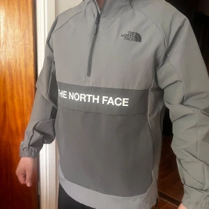 Grå vindjacka från The North Face - Säljer en grå vindjacka från The North Face med huva och halv dragkedja framtill. Jackan har en stor logga över bröstet och elastiska muddar vid ärmsluten. Perfekt för blåsiga dagar och enkel att slänga på sig.