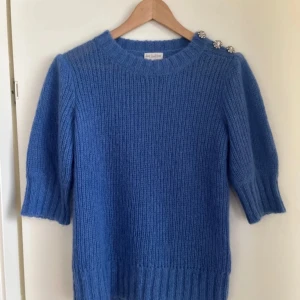 Blå stickad tröja med dekorativa knappar från Dea Kudibal - Mysig blå stickad tröja från Dea Kudibal med tre dekorativa silverknappar på axeln. Tröjan har halvlånga ärmar, nypris 2500kr