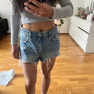 Blå jeansshorts med fransar från Zara - Säljer ett par blå jeansshorts från Zara med råa fransade kanter och slitningar. Shortsen har klassisk femficksmodell, bälteshällor och knappgylf. Perfekta för en avslappnad stil.