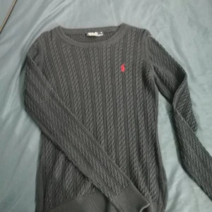 Mörkgrå kabelstickad tröja från Polo Ralph Lauren - Säljer en mörkgrå kabelstickad tröja från Polo Ralph Lauren med klassisk röd logga broderad på bröstet. Tröjan har rund halsringning och långa ärmar, perfekt för lager-på-lager-stil.