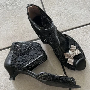 Svarta spetsiga sandaletter med rosett - Svarta sandaletter med låg klack och öppen tå. Skorna har detaljer i spets och en rutmönstrad rosett framtill. Dragkedja baktill för enkel påtagning. Perfekta för dig som vill ha något unikt och snyggt.