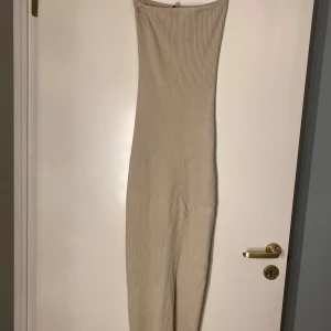 Beige ribbad långklänning - Säljer en beige ribbad långklänning med tight passform. Klänningen är axelbandslös och har en stilren, minimalistisk look som passar perfekt till sommaren. Materialet är stretchigt och följsamt för en bekväm känsla.