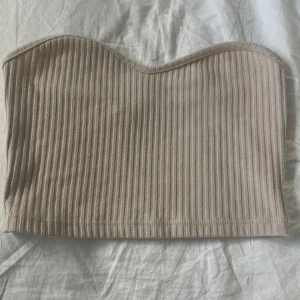 Beige ribbad tubtopp från NLY Trend - Säljer en beige ribbad tubtopp från NLY Trend. Toppen är ärmlös och har en mjuk, stretchig passform som sitter snyggt på kroppen. Perfekt att styla till jeans eller kjol för en enkel och trendig look.