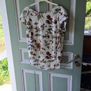 Vit blommig blus - Säljer en vit blus med färgglatt blommönster. Blusen har korta ärmar och rund halsringning. Perfekt för en somrig look och passar till både jeans och kjol.
