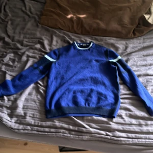 Blå stickad tröja med ränder från Hugo boss  - Säljer en blå stickad tröja från Hugo boss med mörkblå och ljusblå ränder på ärmar och över bröstet. Tröjan har rund halsringning!