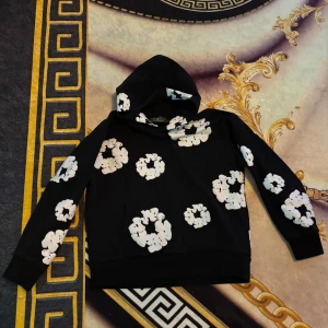 Svart hoodie med vita blommor från Denim Tears - Säljer en svart hoodie från Denim Tears med vita blommönster över hela tröjan. Tröjan har huva och långa ärmar, perfekt för en avslappnad stil. Mjuk och skön med klassisk passform.