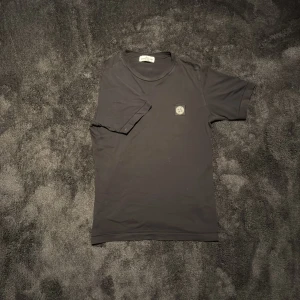 Svart t-shirt från Stone Island  - Säljer en svart t-shirt från Stone Island med klassisk logotyp-patch på bröstet. T-shirten har rund halsringning och är tillverkad i mjuk bomull. Perfekt för en stilren och avslappnad look.