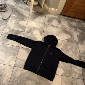 Mörkblå hoodie från Lacoste med dragkedja - Säljer en mörkblå hoodie från Lacoste med dragkedja framtill och klassisk krokodillogga på bröstet. Tröjan har huva och ribbade muddar. Perfekt för en sportig och avslappnad stil. Anledningen varför jag säljer den är för liten 