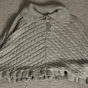 Grå stickad poncho från KappAhl - Superfin grå stickad poncho från KappAhl med kabelstickat mönster, krage och tre knappar framtill. Volangkant nedtill ger en extra söt touch. Perfekt att slänga över en tröja när det är kyligt ute.🥰🥰💕