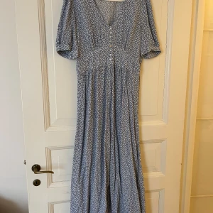 Blommig långklänning från Indiska - Söt blå långklänning med vitt blommönster från Indiska. Klänningen har v-ringning, knappar framtill och korta ärmar. Perfekt för sommarens alla tillfällen. Lätt och luftig modell med smock i ryggen för extra komfort.