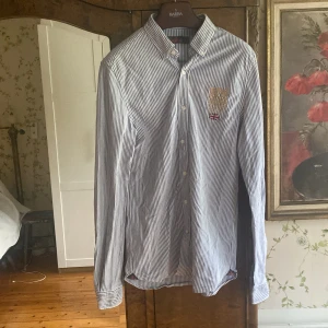 Morris skjorta - Snygg blå och vit randig skjorta från Morris med broderad logga och brittisk flagga på bröstet. Skjortan har klassisk krage med knappar och långa ärmar. Perfekt för en stilren look. Perfekt för sommaren. 