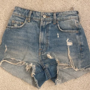 Blå jeansshorts med slitningar från ZARA - Säljer ett par blå jeansshorts från ZARA med hög midja och råa, fransiga kanter. Shortsen har flera slitningar och en klassisk femficksdesign. Perfekta för en avslappnad stil.