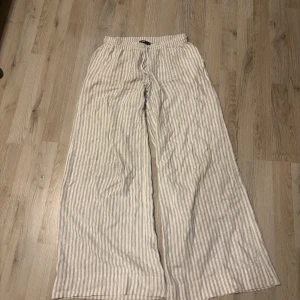 Randiga linnebyxor från BikBok - Säljer ett par vita linnebyxor med tunna grå ränder från BikBok. Byxorna har resår och snörning i midjan samt vida ben för en avslappnad look. Perfekta för varma dagar.