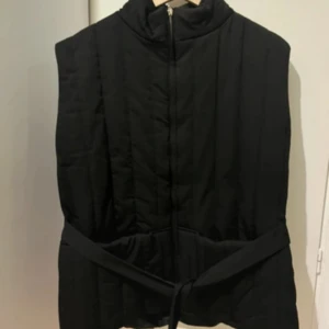 Svart quiltad väst med bälte - Snygg svart quiltad väst med hög krage och knytbälte i midjan. Perfekt att slänga över en hoodie eller tröja för extra värme och stil. Västen har dragkedja framtill och en enkel, stilren design.