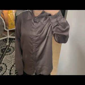 Grå satinblus med knappar - Snygg grå satinblus med klassisk krage och knäppning framtill. Långa ärmar med breda manschetter och en lätt glansig finish som ger en lyxig känsla. Perfekt att styla till olika looks.