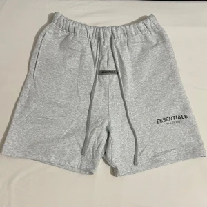 Grå shorts från Essentials Fear of God - Ljusgrå shorts från Essentials Fear of God med elastisk midja och snörning. Mjukt material och logga framtill. Perfekta för en avslappnad stil.